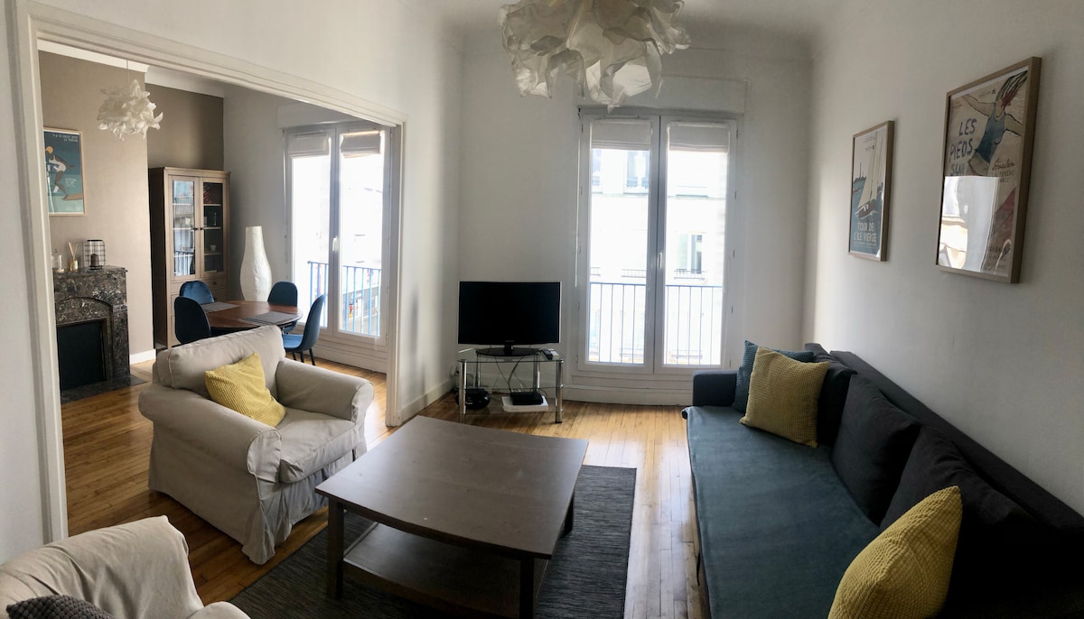 A spacious living room is presented, featuring a large coffee table at its center. Comfortable sofas with decorative cushions are positioned around the room. A television sits against one wall, while large windows allow natural light to illuminate the area. A dining space is visible through an adjoining doorway.