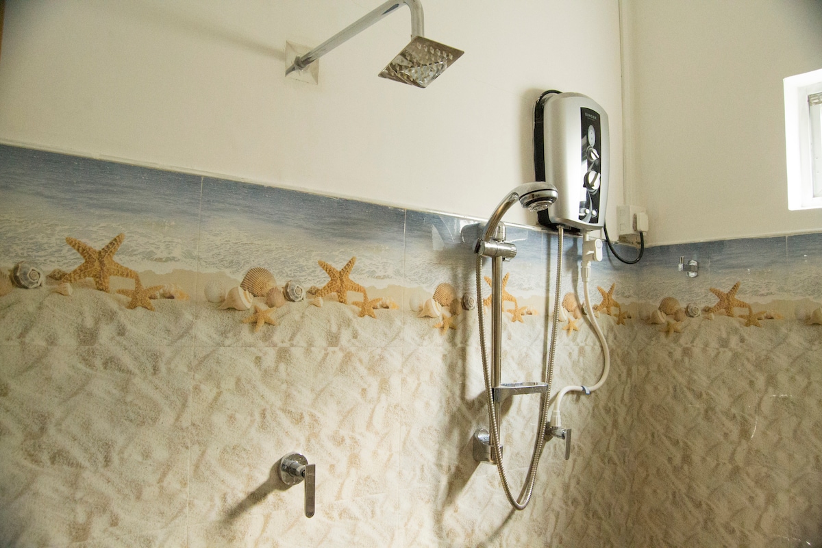 The bathroom features a tiled wall adorned with a seashell motif, including starfish and shells. A modern showerhead and a water heater are installed, with sleek fixtures providing a clean and functional design. Natural light enters through a small window.