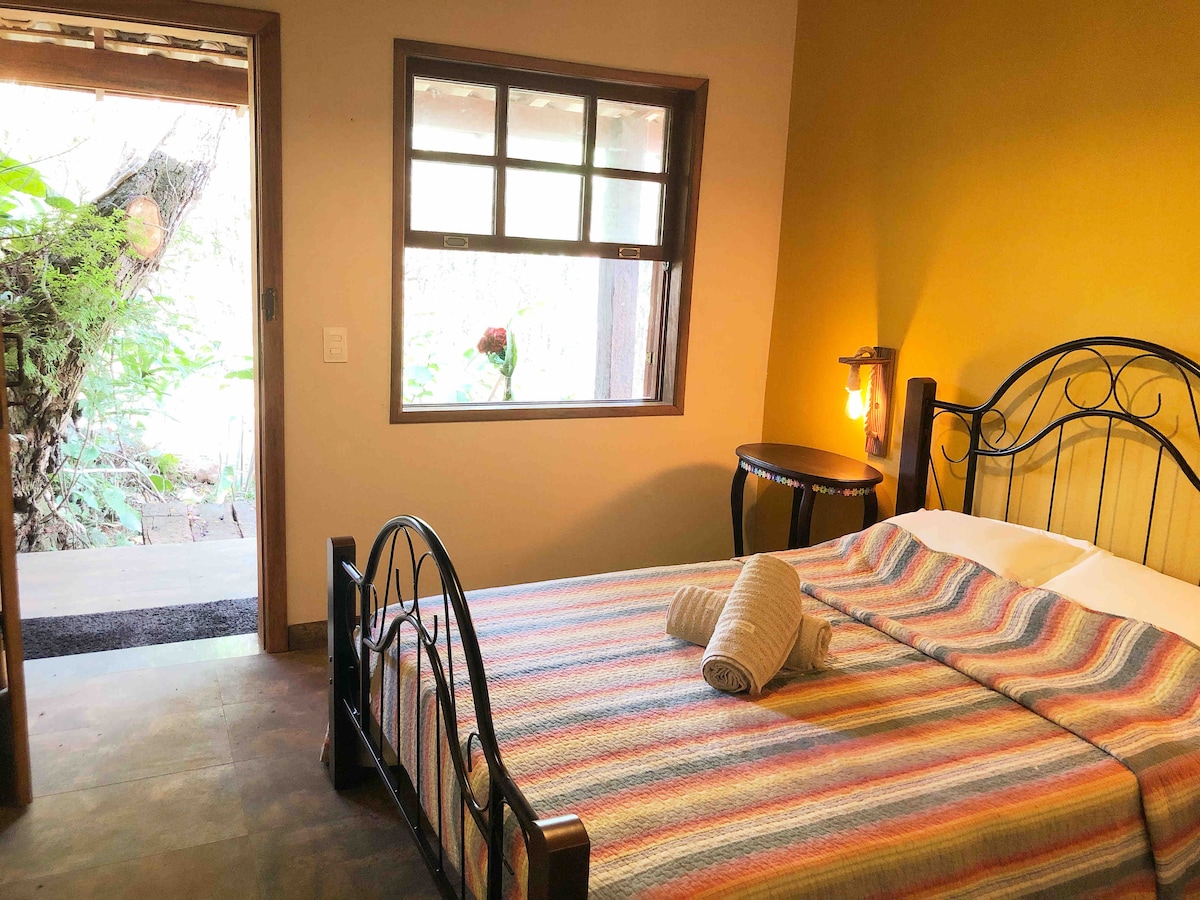 A cozy sleeping area features a striped blanket over a spacious bed, complemented by a wooden bed frame. Natural light filters in through a window, illuminating the room. A small table sits nearby with a towel neatly arranged on it, adding a touch of simplicity.