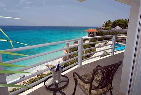 A balcony view showcases a sparkling ocean in various shades of blue. A small table holds a champagne bucket, and a comfortable chair is positioned to appreciate the scenery. In the background, a pool and palm trees contribute to the coastal ambiance.