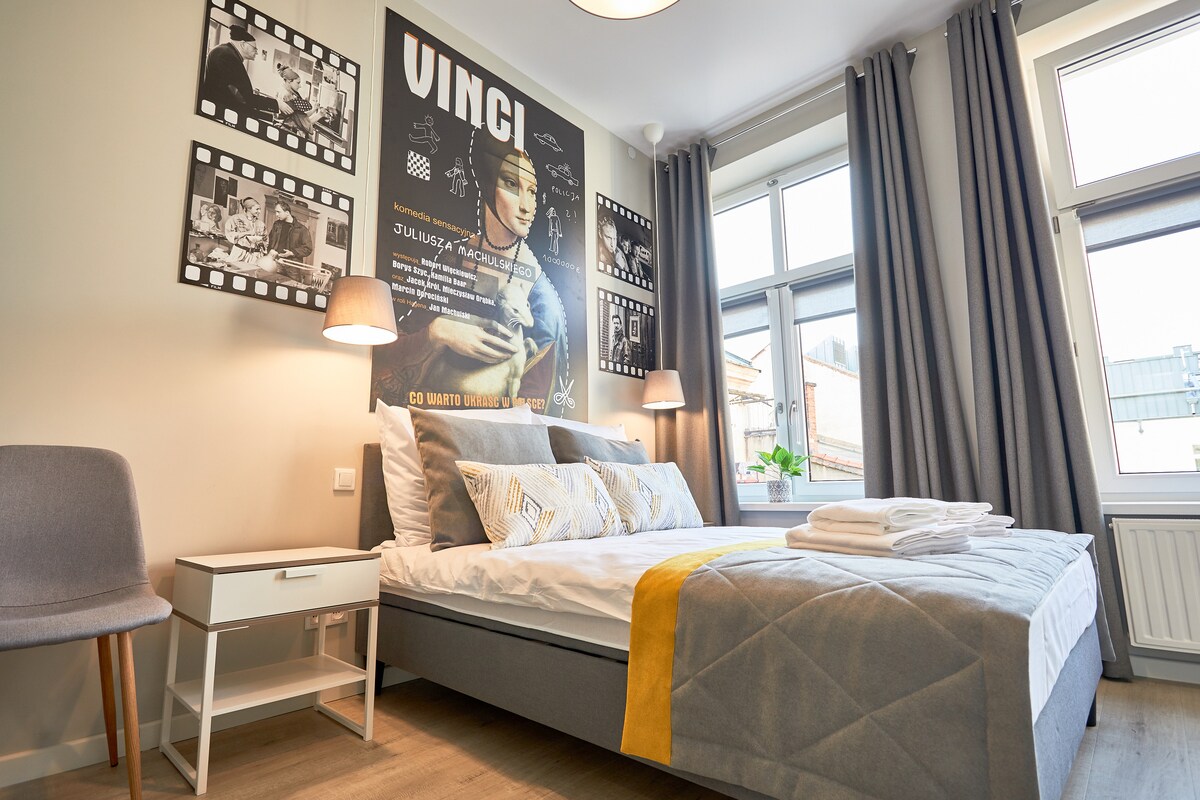 A bright sleeping area is highlighted by a large mural featuring artistic film-themed elements. The spacious bed is dressed in crisp white linens with a yellow accent. Two bedside lamps softly illuminate the space, complemented by neatly arranged towels on the bed. Two large windows allow natural light to enter.