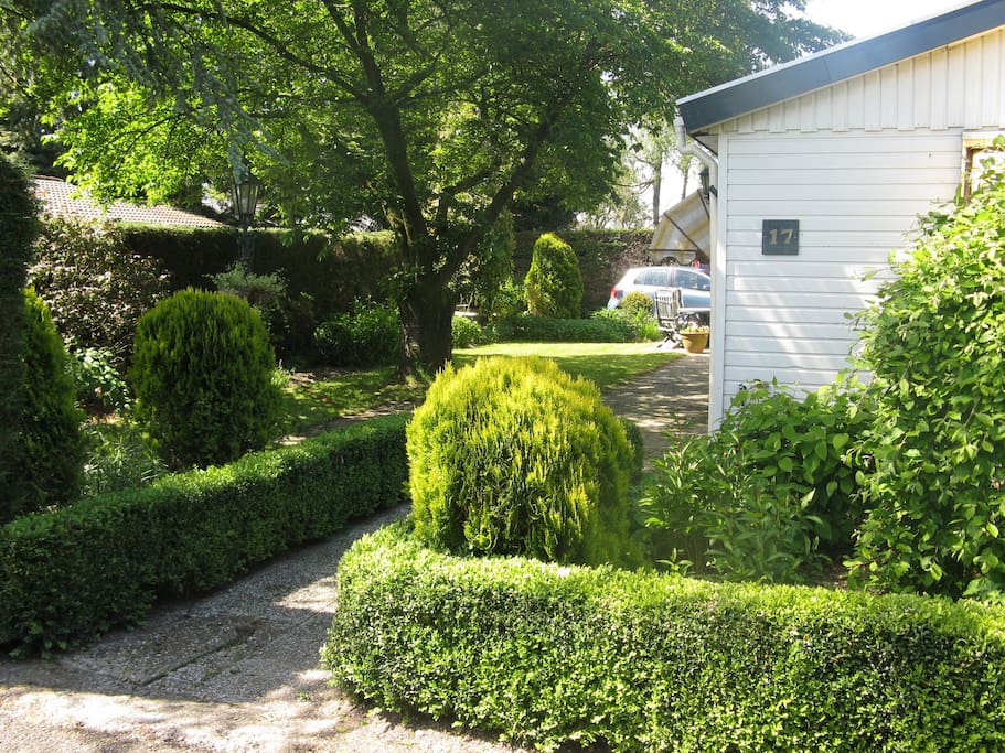 Voorzijde bungalow. Tuin met veel privacy. Er zijn 4 verstelbare tuinstoelen aanwezig en op de veranda staan nog twee extra stoelen. Aan de acherzijde is de oprit voor de auto, inclusief carport.