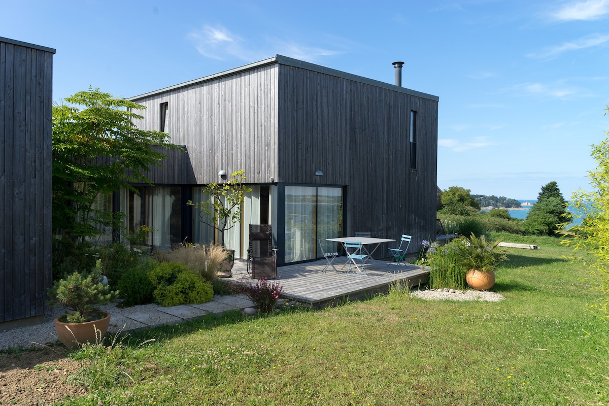 The exterior of a modern wooden house is visible, featuring large glass doors and a deck with a table and chairs. Lush greenery surrounds the house, while the serene bay is seen in the distance, complemented by a blue sky.