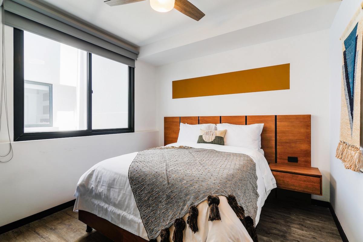A well-lit bedroom features a spacious bed with a textured throw draped across the foot. An accent wall in warm tones complements the wooden headboard, while natural light filters through large windows covered by sleek blinds.