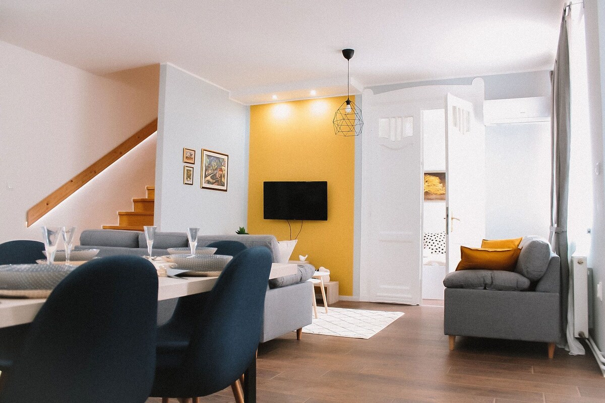 An open living space features a dining area with a light wooden table and blue upholstered chairs. A soft gray sofa complements the room, which is illuminated by natural light. A staircase can be seen in the background, leading to the upper level.