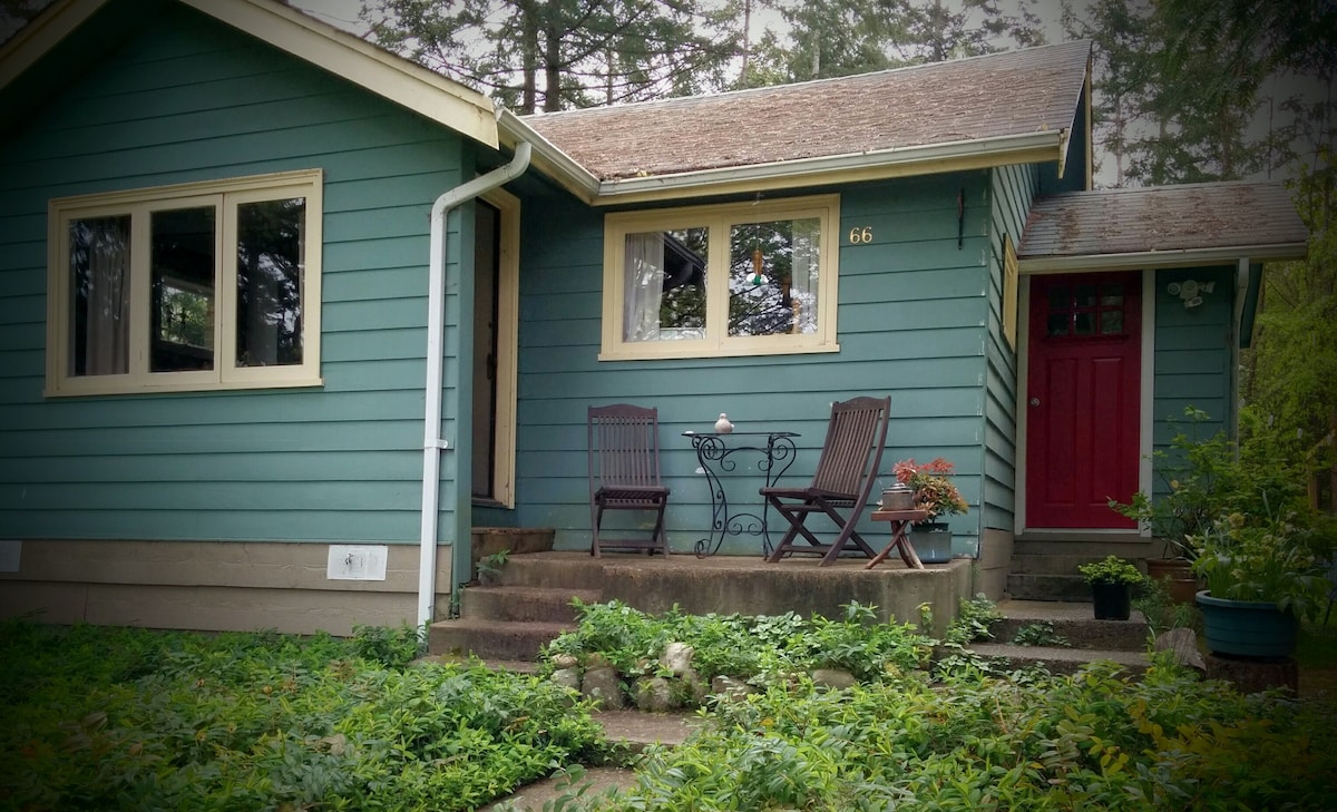 The cottage exterior features a blue-green facade with a red front door. A small patio area is visible, containing two wooden chairs and a table adorned with potted plants. Steps lead up from the lush garden, which surrounds the entrance.