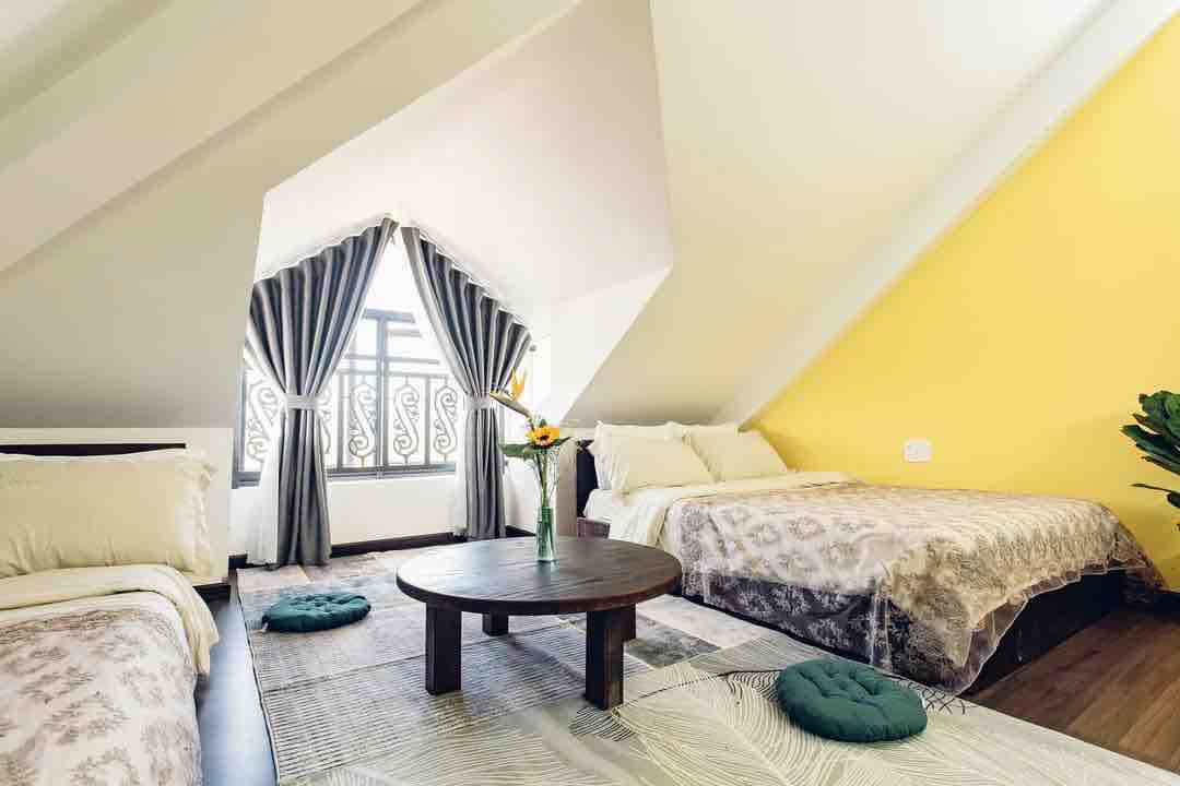 A brightly lit room features two spacious beds adorned with soft blankets. Natural light filters through large curtains framing a geometric-patterned window. A round wooden table is centered on a textured rug, accompanied by green floor cushions. One wall is painted in a cheerful yellow.