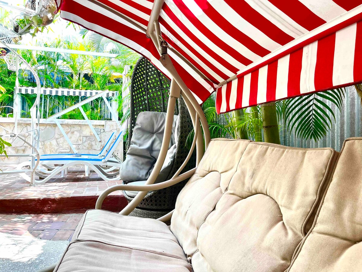 A shaded seating area is featured with a comfortable cushioned sofa beneath a striped canopy. Surrounding plants add greenery to the scene, while a lounge chair is visible nearby, enhancing the inviting atmosphere of the outdoor space.