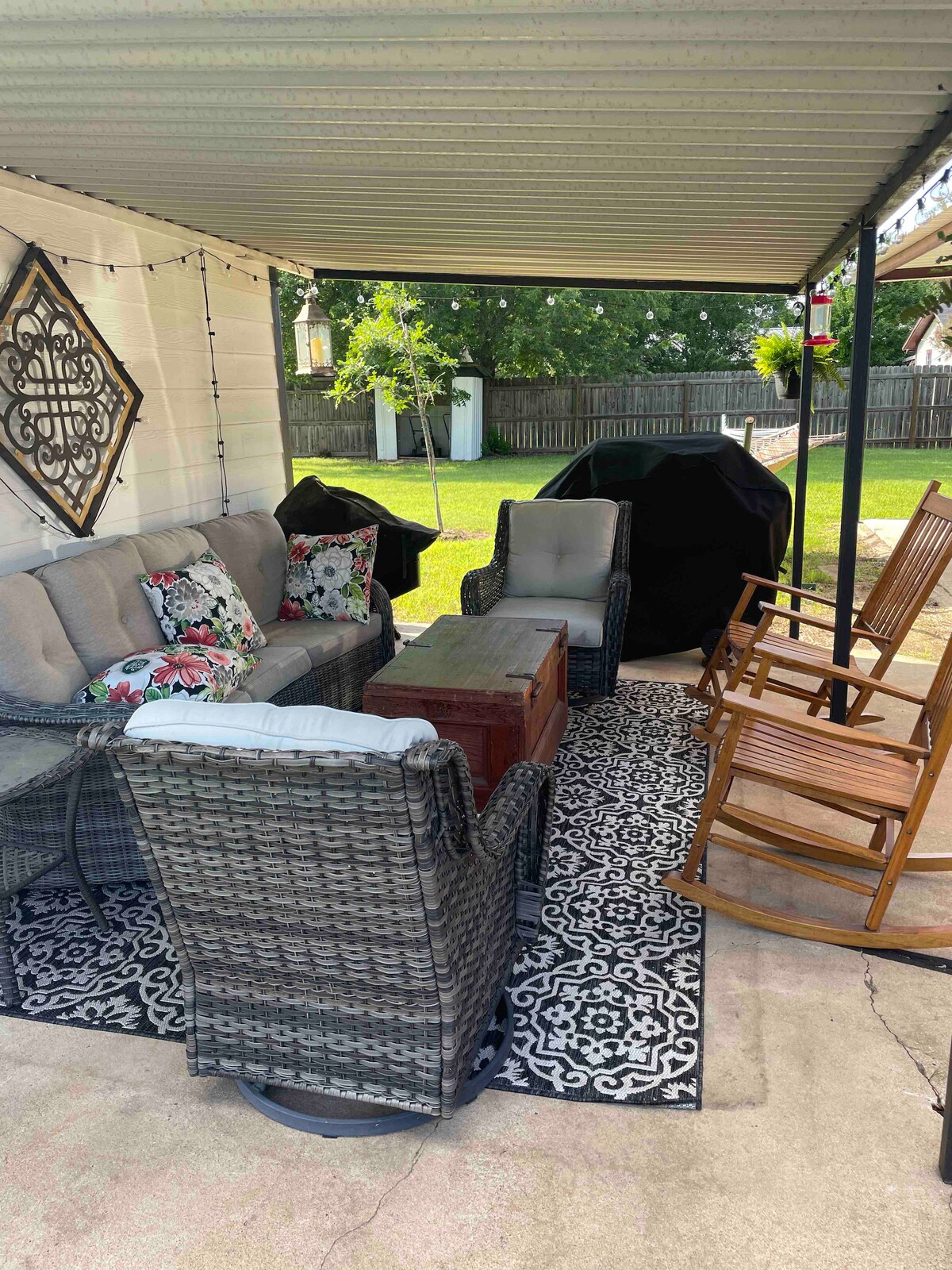 An outdoor seating area features a comfortable arrangement of furniture, including a gray sofa adorned with floral cushions and two chairs. A wooden coffee table sits at the center, complemented by a patterned rug beneath. Rocking chairs are positioned nearby, with a spacious green yard visible in the background.