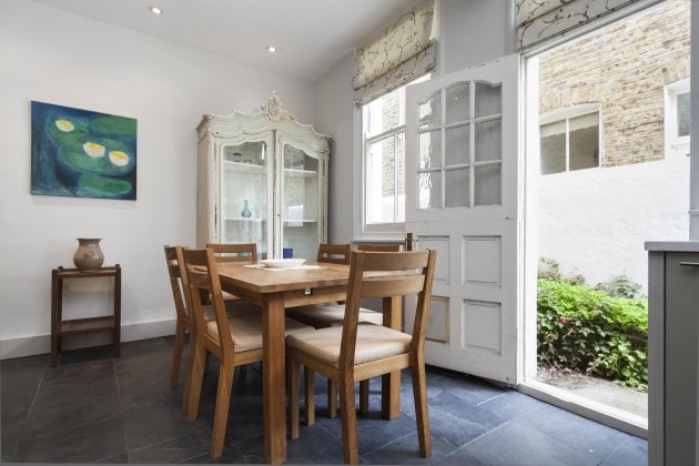 A well-lit dining area features a wooden table surrounded by six chairs. A light-colored cabinet is positioned against the wall, showcasing decorative items. Natural light streams in through the door leading to a small garden, enhancing the space's brightness.
