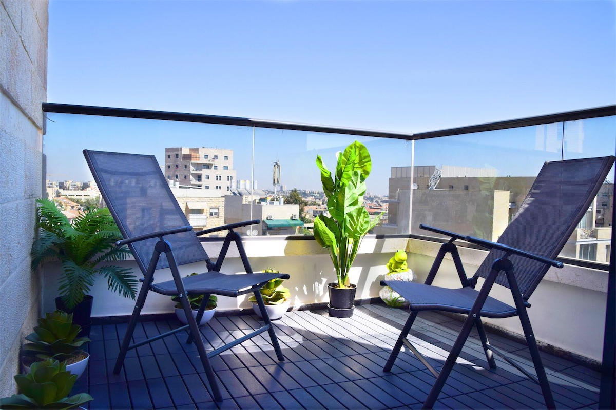 A modern terrace is showcased with two black lounge chairs positioned around a potted plant. The wooden flooring contrasts with the green plants nearby, while the surrounding glass railing offers unobstructed views of the city skyline under a clear blue sky.