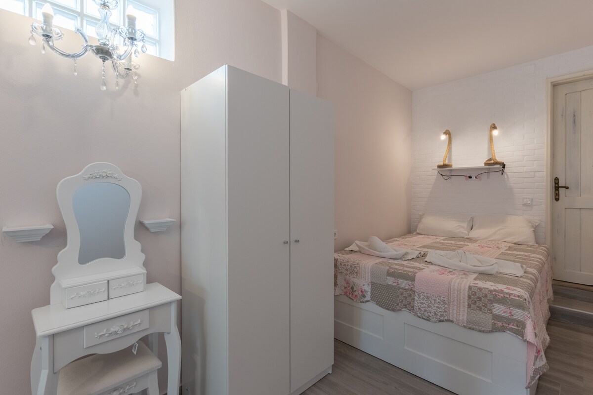 A softly lit bedroom features a spacious bed with neutral-toned bedding. A white wardrobe with clean lines stands next to a vintage-style vanity with a mirror. Wall-mounted lamps illuminate the space, while light from a small window adds warmth to the pastel walls.