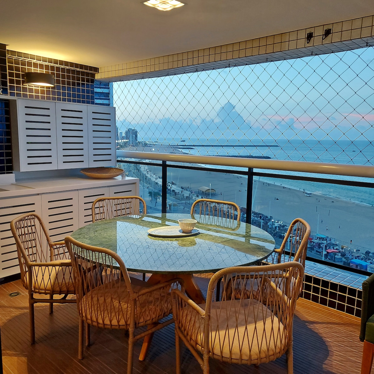 A dining space is featured with a round table surrounded by four rattan chairs, set against a backdrop of a wide ocean view. Large windows provide natural light and the horizon is visible, enhancing the seaside dining experience.