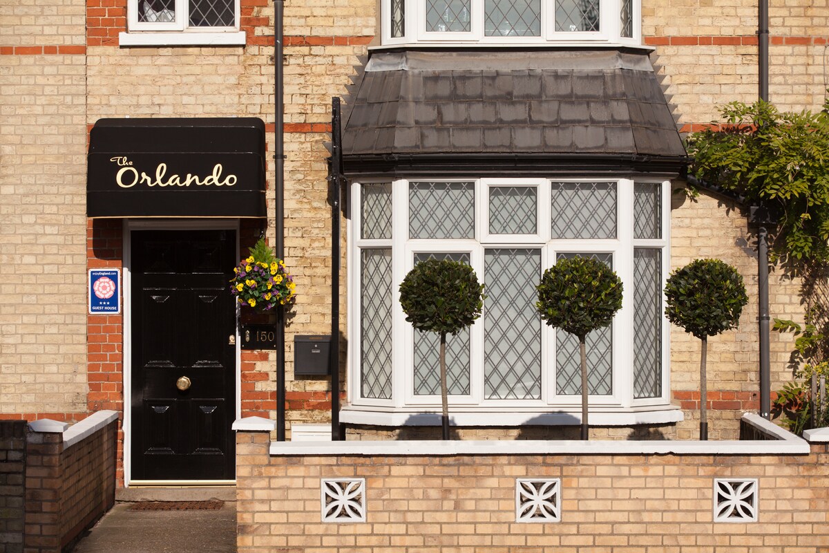 The entrance of The Orlando guest house features a stylish black door with a circular handle and the name displayed above. Flanking the entrance are two neatly trimmed trees in pots, with a decorative flower arrangement adding a touch of color. The brick façade is well-maintained, enhancing the inviting look.