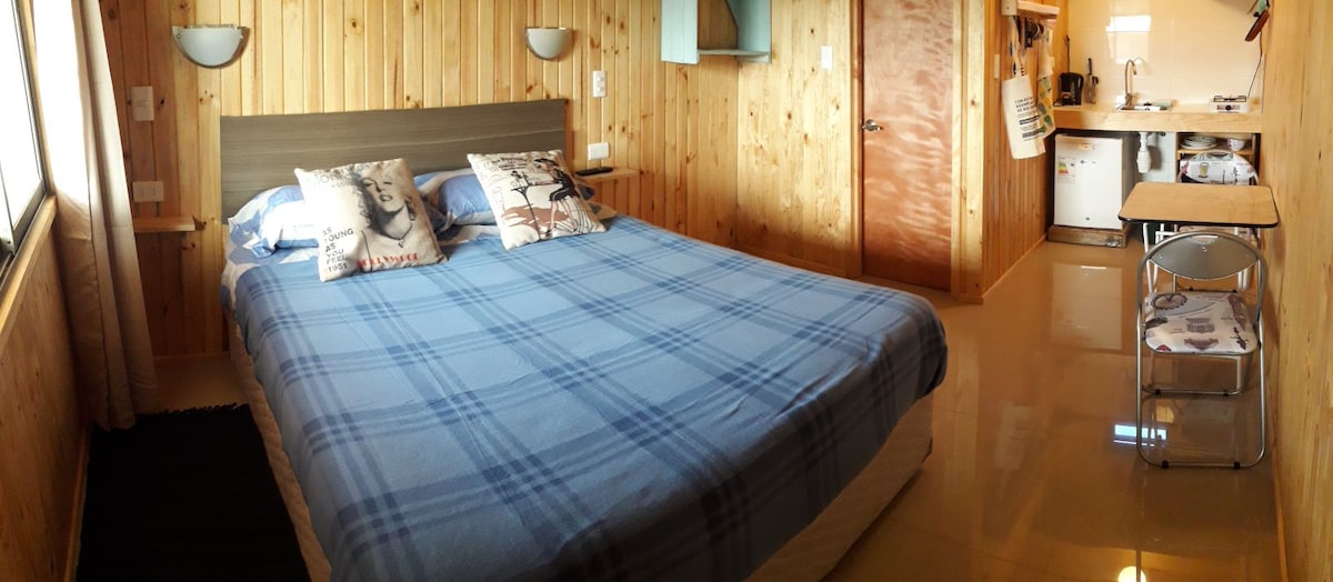 A wooden-paneled mini apartment features a spacious bed adorned with a plaid blanket. Soft lighting from wall sconces illuminates the room, which includes a kitchenette visible in the background. A small dining table with chairs is positioned near the entrance, creating a functional living space.