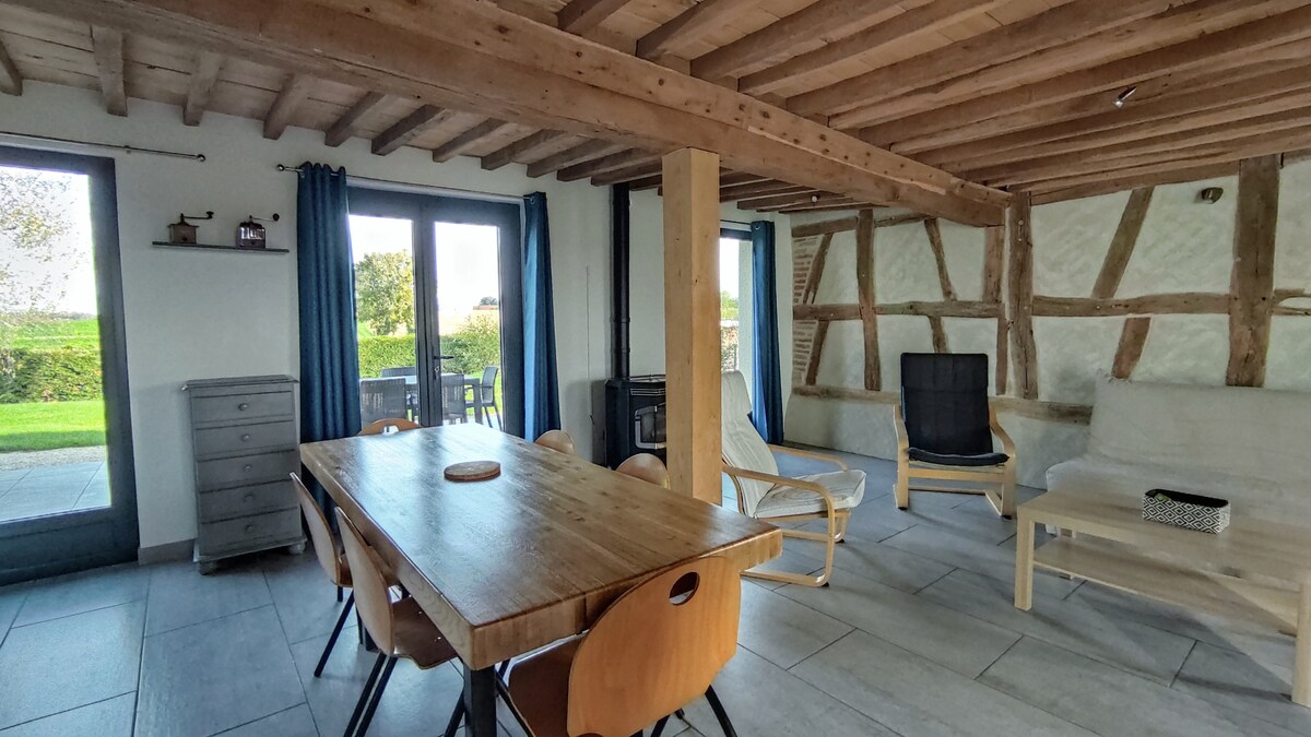 A spacious living area features a rustic wooden ceiling and large windows that provide natural light and a view of the outdoor greenery. A wooden dining table is surrounded by modern chairs, while a comfortable seating arrangement includes a chaise lounge and a black armchair.