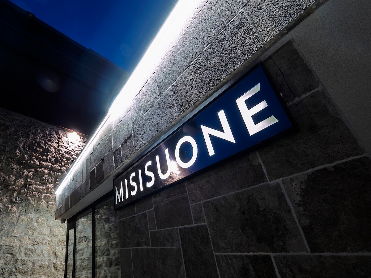 The exterior of the Misisuone apartments is showcased, featuring a prominent illuminated sign against a stone wall. The light creates a welcoming ambiance, highlighting the modern architectural details and providing clear visibility of the establishment's name.