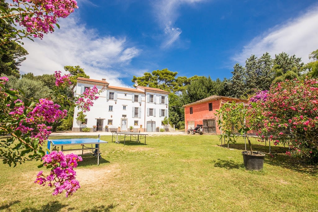 The Villa is framed by vibrant flowering plants and greenery, showcasing a spacious garden area. Two buildings, the main Villa and a red structure, are visible against a clear blue sky. Outdoor tables suggest opportunities for leisure and gatherings in this peaceful, natural setting.