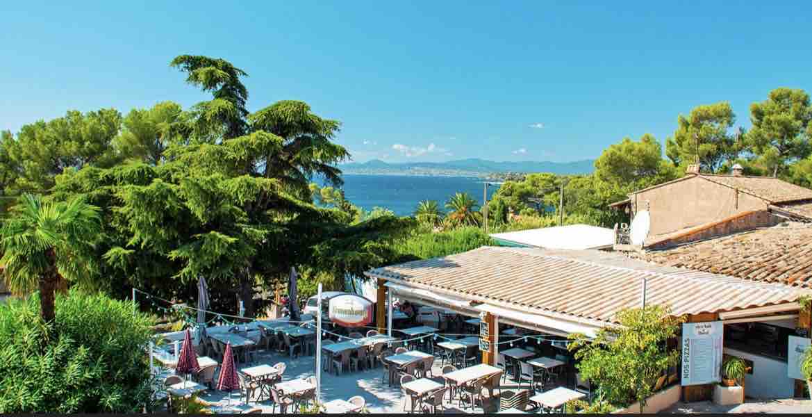 An outdoor dining area is shown under a clear blue sky, surrounded by lush greenery and palm trees. Tables and chairs are arranged neatly, with views of the sea and distant hills visible in the background.
