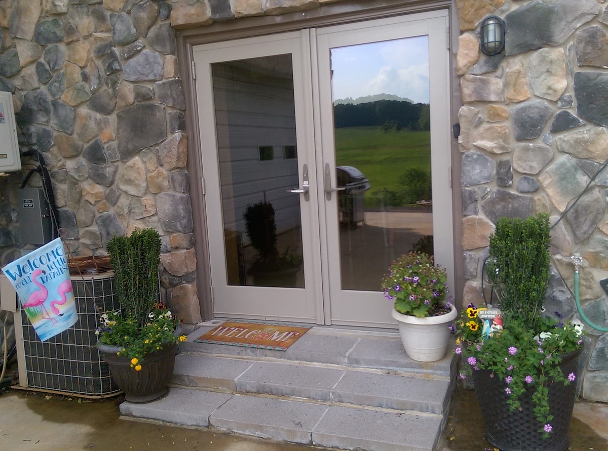 The entrance features a pair of glass doors framed by a stone exterior, flanked by potted plants with colorful blooms. A welcome mat is positioned on the stone steps, enhancing the inviting nature of the entryway. A banner is displayed nearby, adding to the warm surroundings.
