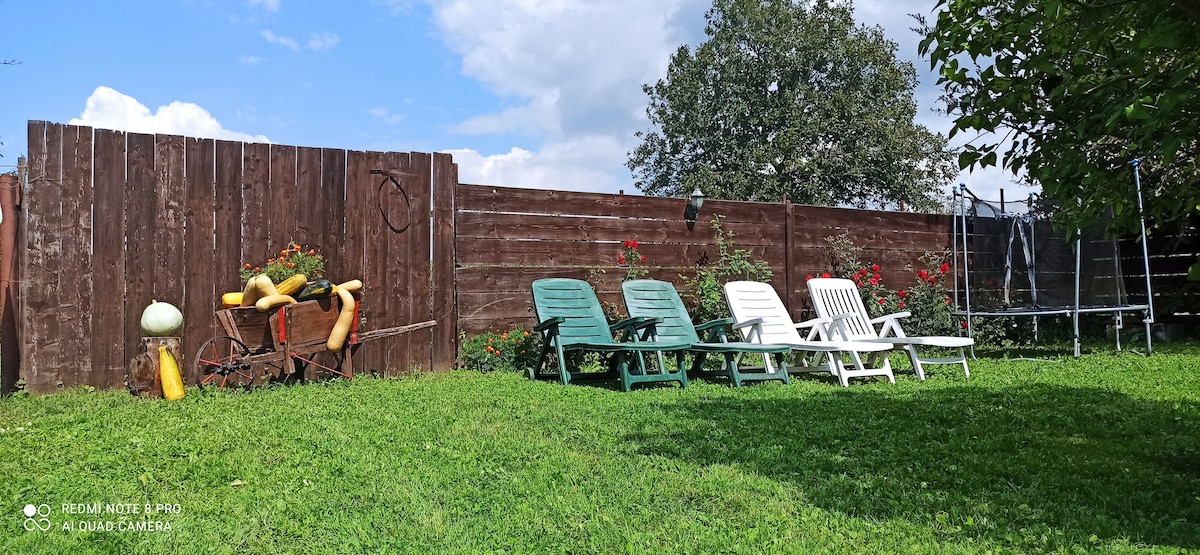 A spacious backyard is depicted with a green lawn, featuring several reclining chairs in varying colors along the wooden fence. A row of vibrant flowers adds color to the scene, while a trampoline can be seen in the background, inviting outdoor activities.