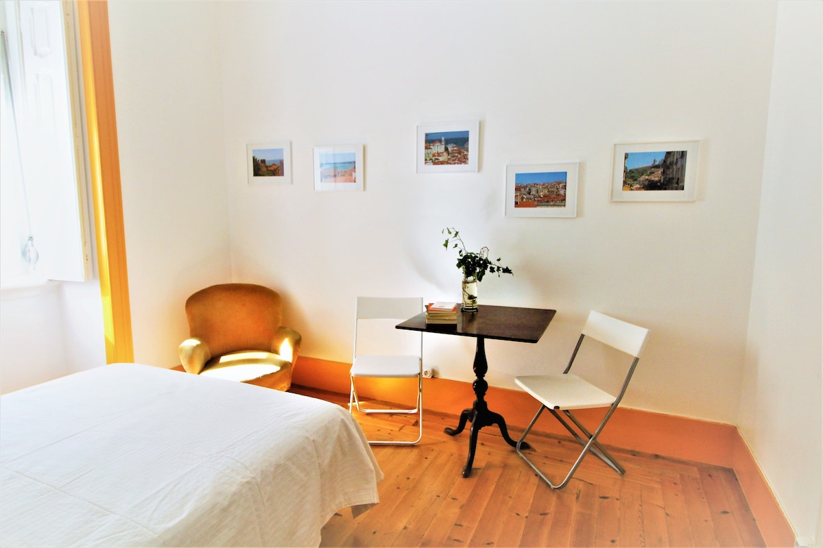 The studio features a spacious bed in the foreground, complemented by a small wooden table and two folding chairs. A cozy armchair sits nearby, and the wall displays a series of framed photographs. Natural light enters through a large window, illuminating the warm wooden floor.