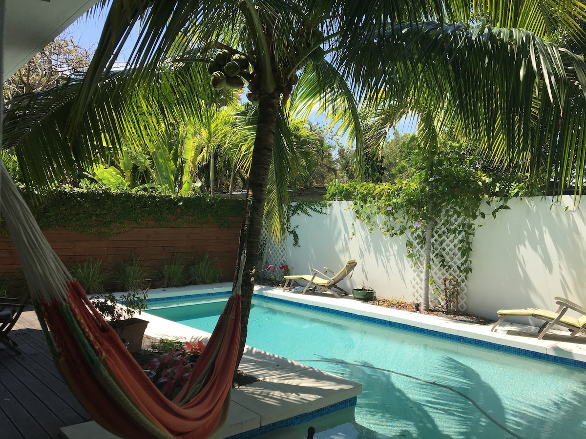 The image features a serene outdoor area with a swimming pool surrounded by lush greenery. A hammock is seen hung between palm trees, with sun loungers positioned near the water's edge. The atmosphere is calm, enhanced by the vibrant tropical plants lining the pool.