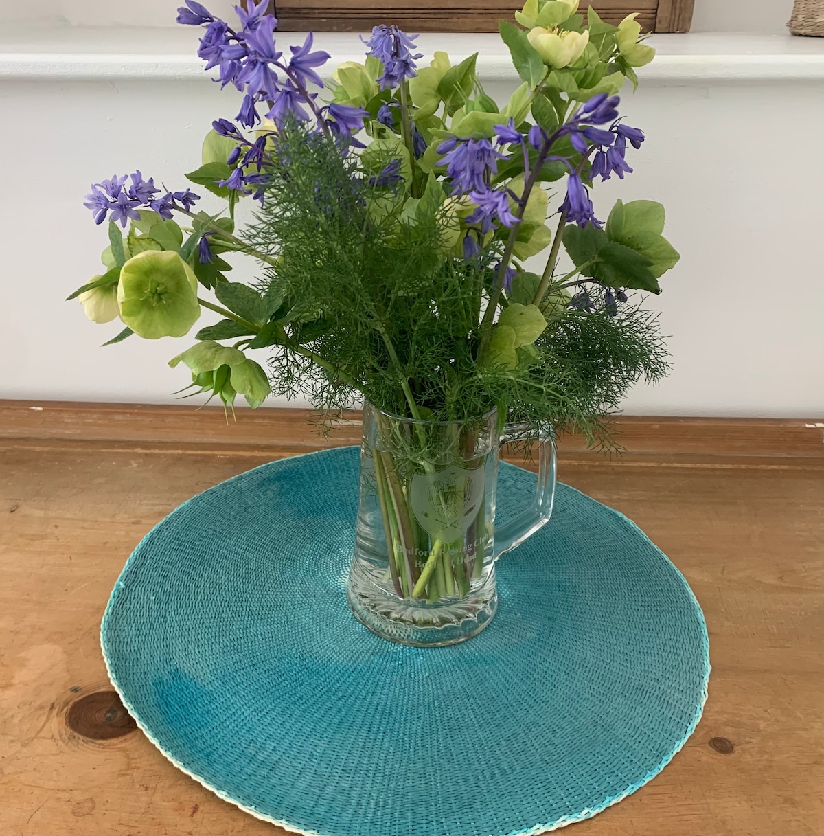 A clear glass vase filled with purple and green flowers sits atop a round turquoise placemat. The arrangement features delicate foliage, providing a fresh and vibrant touch to the wooden surface underneath.