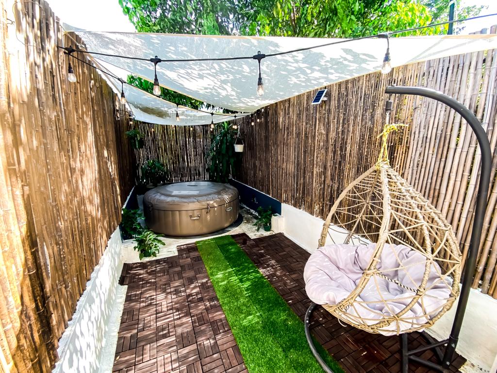 A private outdoor space features a hot tub surrounded by bamboo fencing and greenery. A hanging chair is placed on a wooden deck, with an artificial grass pathway leading through the area. Decorative string lights are draped above, enhancing the ambiance.