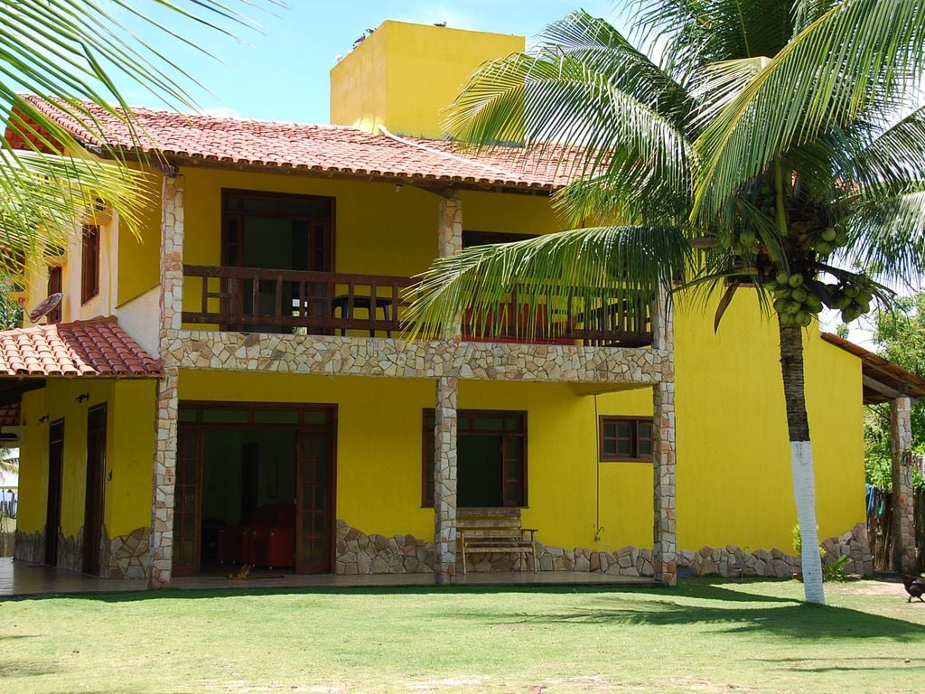 The exterior of the house features vibrant yellow walls and stone accents, flanked by palm trees. A spacious porch is presented with wooden railings, providing a relaxed area for seating. Large windows allow natural light to illuminate the interior, creating an inviting appearance.