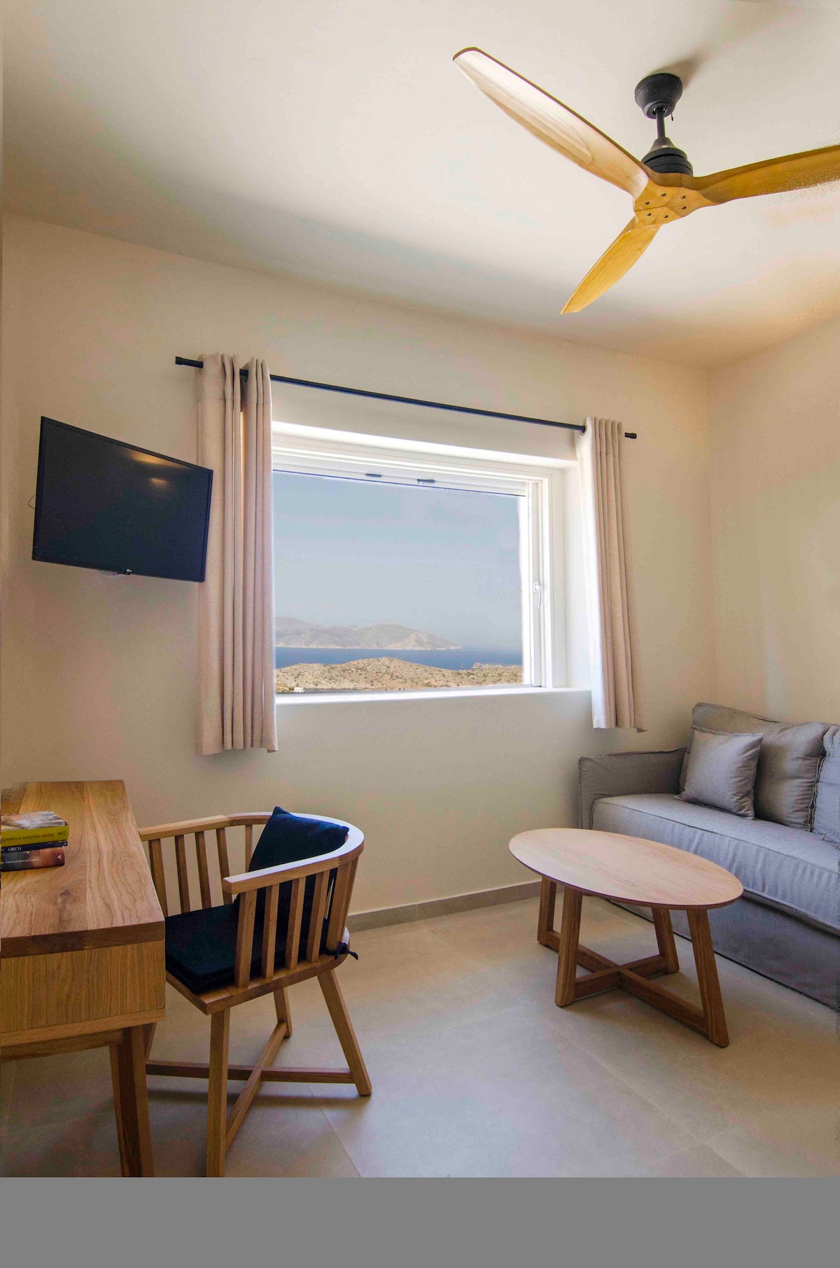 A light-filled living area features a sofa and a small round table, complemented by a wooden desk and a chair. A flat-screen TV is mounted on the wall, and a window offers unobstructed views of the surrounding landscape and sea.