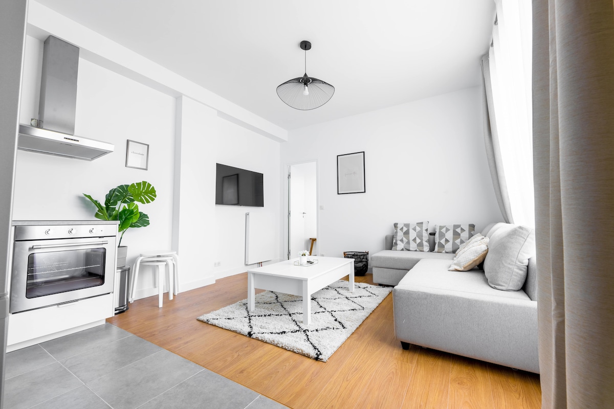 A contemporary living area features a spacious light grey sofa and a modern coffee table centered on a patterned rug. A mounted television is visible on the wall, alongside a kitchenette with stainless steel appliances. Large windows allow natural light to fill the room.
