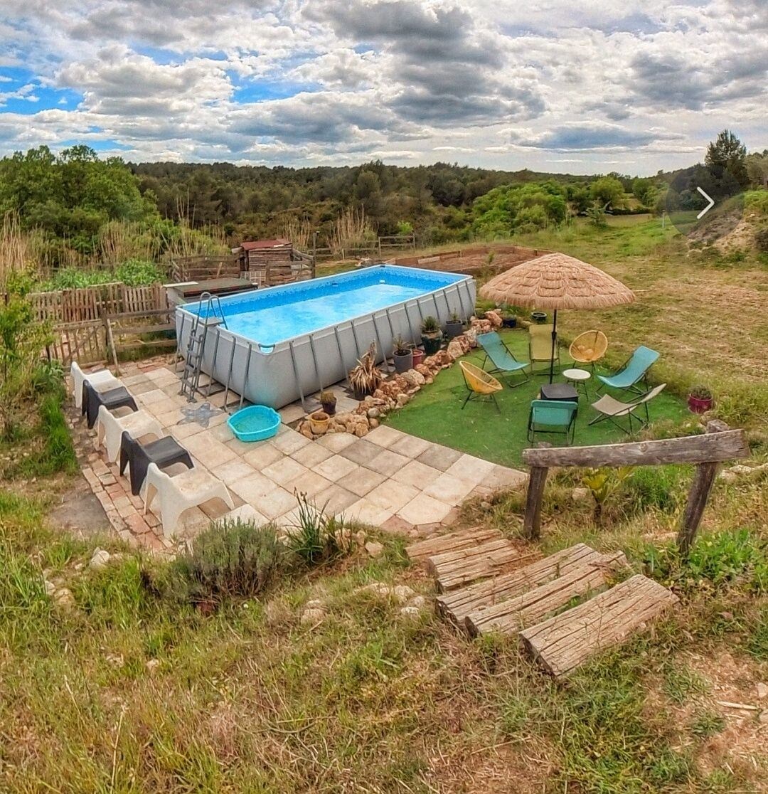 An above-ground pool is surrounded by a stone patio, featuring lounge chairs and a shaded seating area. A grassy expanse connects to natural surroundings, with trees and gentle hills in the background. A small set of wooden steps leads down from the patio.
