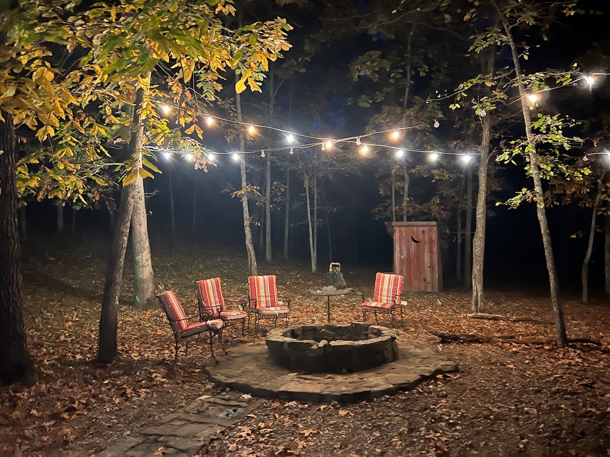 An outdoor fire pit area is surrounded by several striped chairs, all set beneath warm string lights. Trees with autumn leaves create a serene backdrop, enhancing the secluded atmosphere. An outbuilding is visible in the distance, completing the tranquil setting.