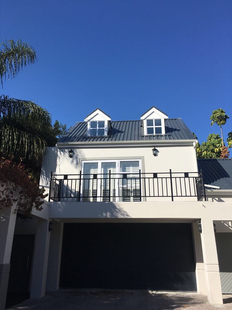 The exterior of a two-story house features a sleek black roof with dormer windows and a balcony. The façade is painted in a light color, complemented by a subtle landscape with greenery and a clear blue sky in the background.