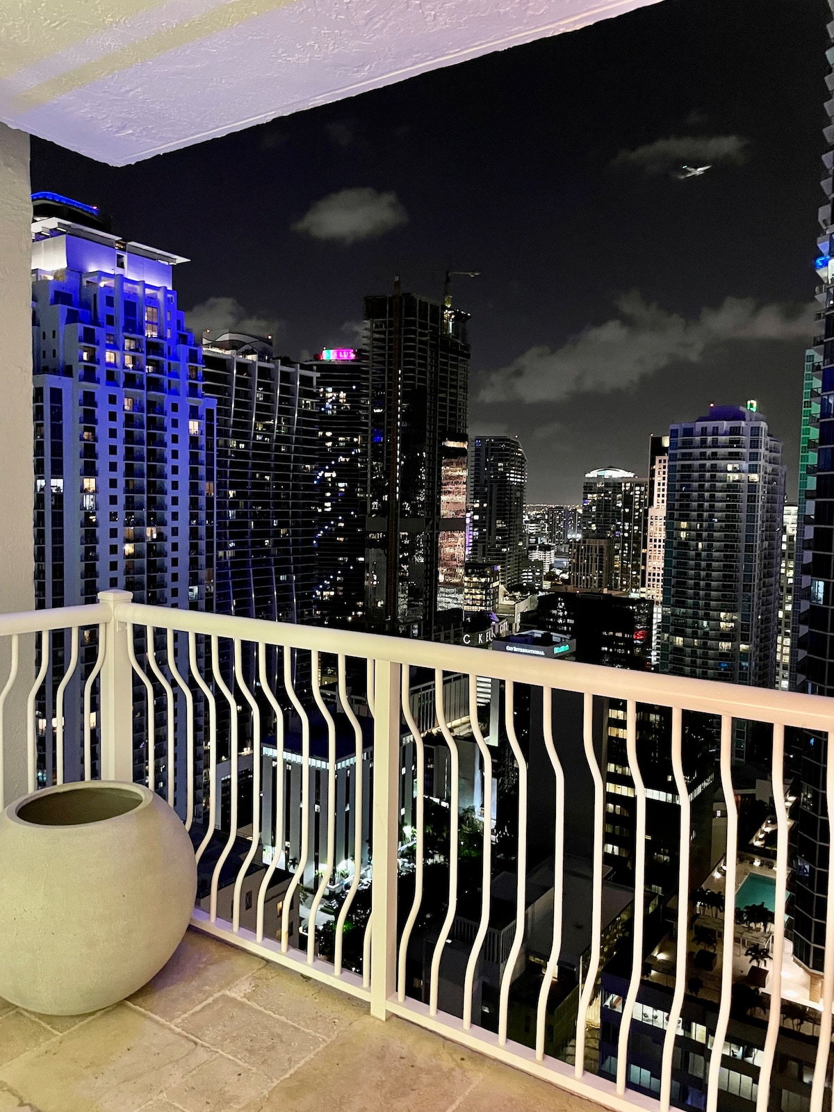 A spacious balcony offers a view of the city skyline at night, with illuminated skyscrapers and vibrant lights. A simple gray planter is positioned on the balcony floor, adding to the modern aesthetic, while the railing provides a sense of openness to the bustling city below.