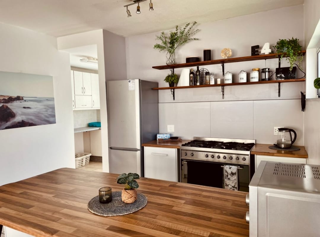 The kitchen features modern cabinetry with open shelving displaying decorative items and kitchen essentials. A large island with a wooden countertop offers ample preparation space. Stainless steel appliances, including a fridge and oven, are present, with natural light illuminating the area from a nearby window.