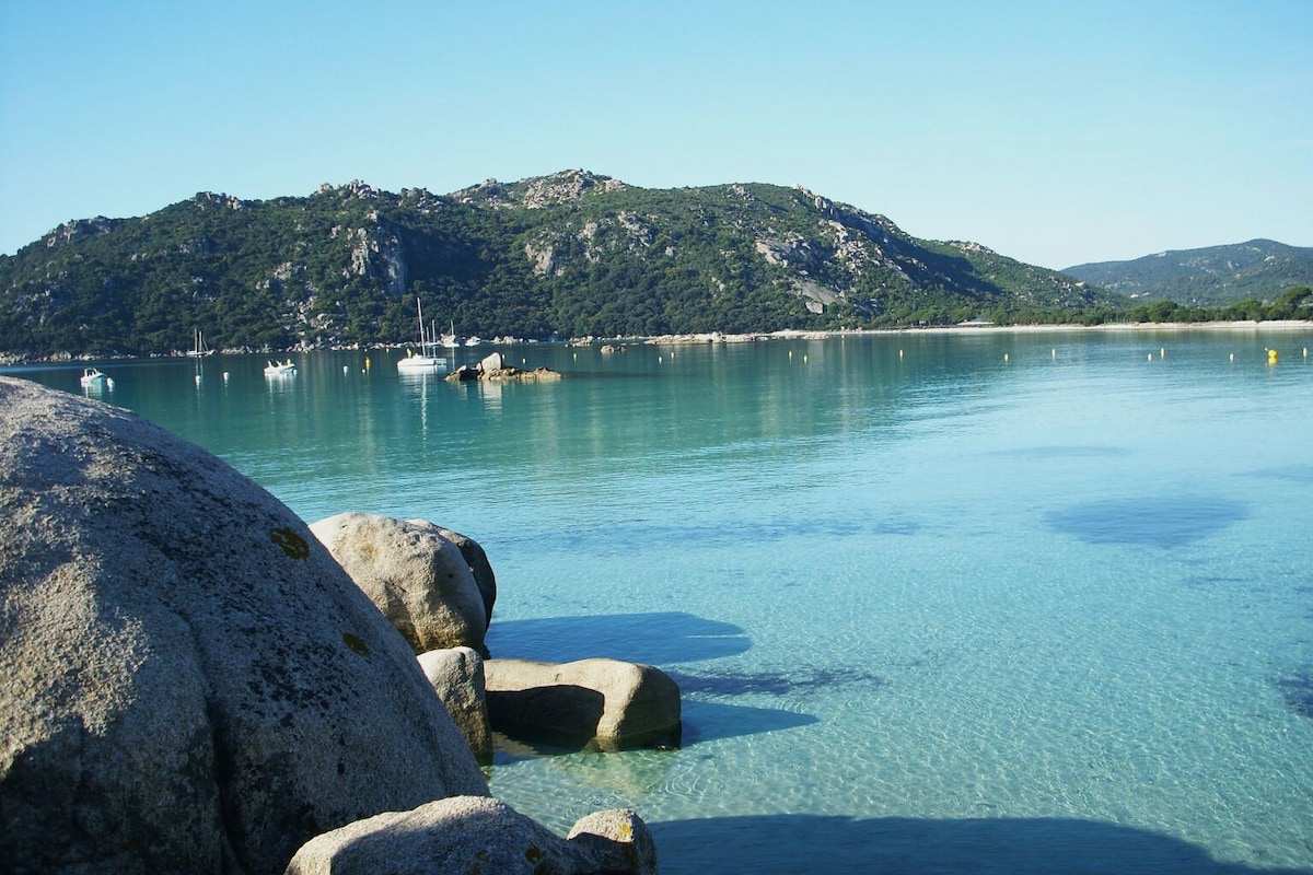Calm waters are depicted, reflecting the clear blue sky, with rolling hills in the background. Smooth, large rocks sit at the water's edge, while boats are anchored in the distance. This serene landscape invites a sense of tranquility.