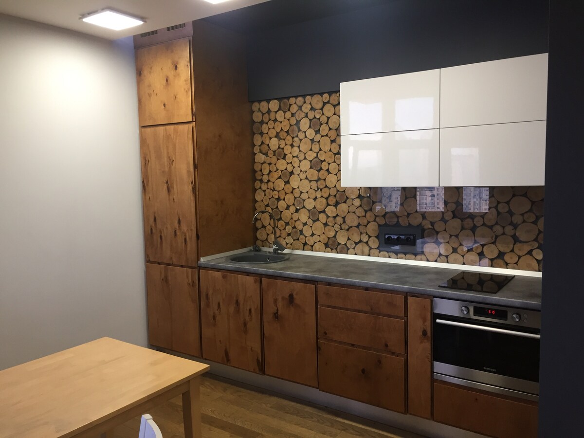 The kitchen features wooden cabinetry accented by a stacked log wall design. A minimalist countertop is complemented by modern appliances, including an oven and stovetop. Ample natural light illuminates the space, which includes a small dining table positioned nearby.