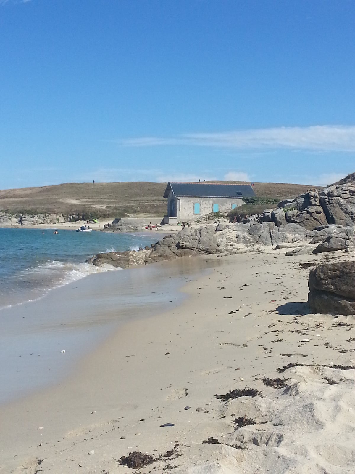 A beach scene features soft, sandy shores leading to calm, clear waters. A small, rustic building is visible on a distant hillside, surrounded by grassy terrain. Large, smooth rocks line the beach, adding texture to the landscape under a bright blue sky.