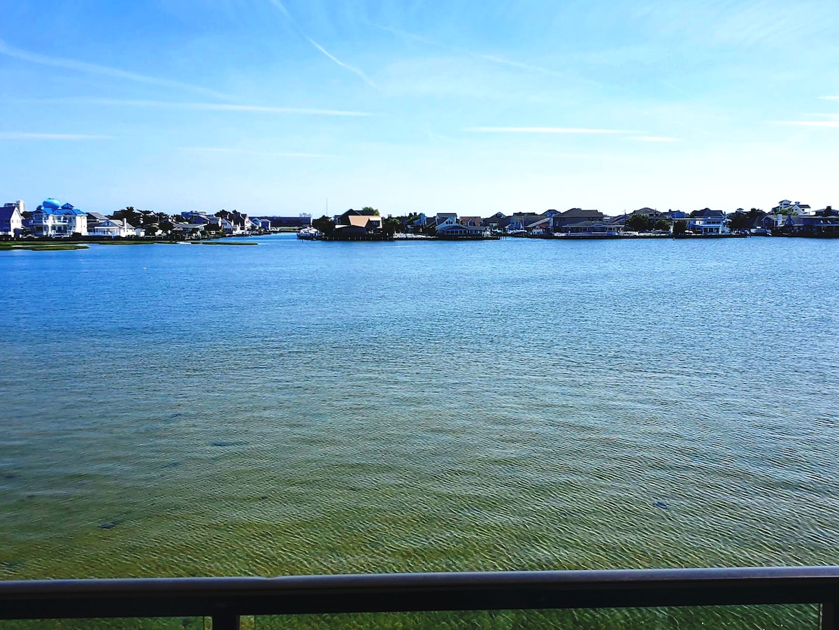 A clear view of the tranquil water extends from the balcony, showcasing vibrant shades of blue and green. Coastal homes are visible along the shoreline, framed by a bright blue sky with wispy clouds.