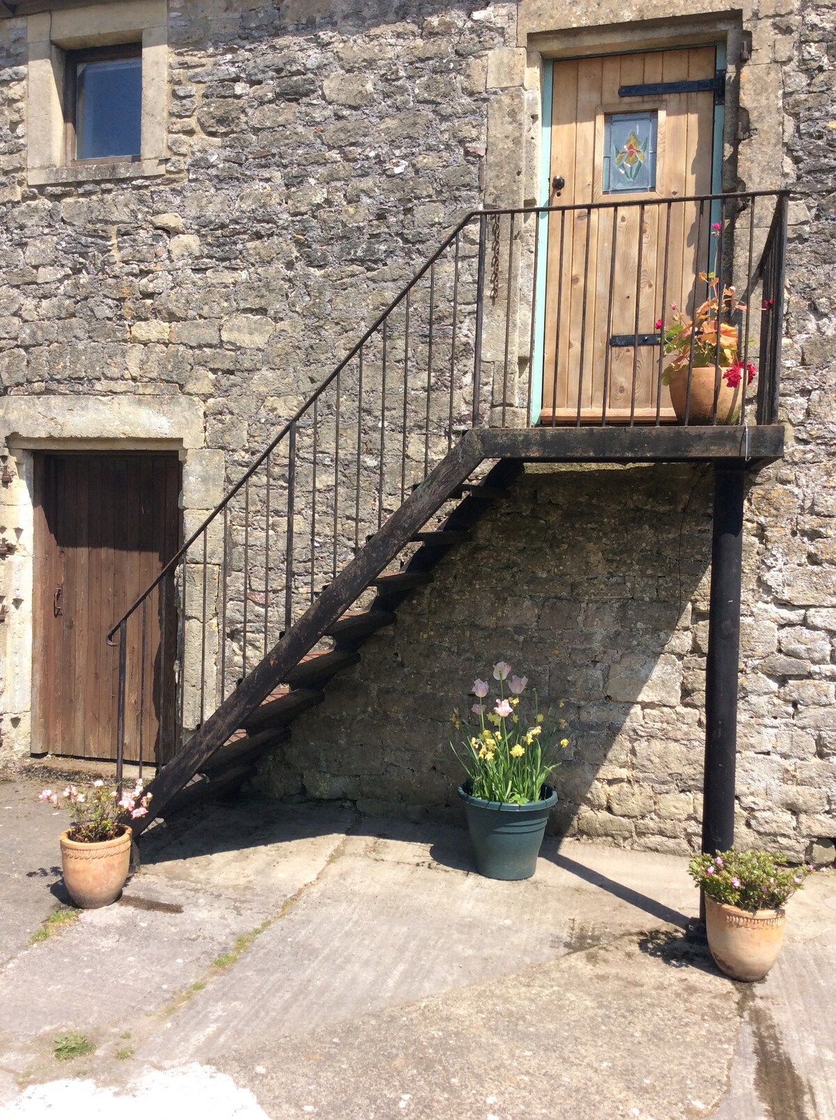 An iron staircase leads to the upper floor studio, surrounded by rustic stone walls. Potted plants adorn the entrance, with colorful flowers adding charm. A wooden door featuring a decorative panel is positioned above the staircase, while a smaller door is visible alongside.
