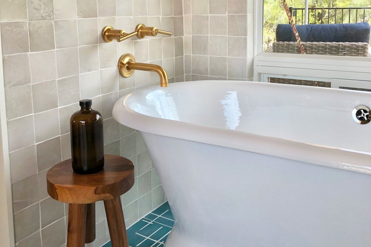 A modern bathroom features a luxurious freestanding tub with smooth, white porcelain. Gold fixtures provide an elegant accent against light-colored tiled walls. A wooden stool sits beside the tub, holding a dark glass bottle, contributing to a well-coordinated design.