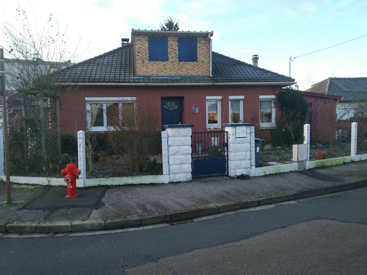 A welcoming single-story house features a red exterior with yellow brick accents above the front door. A white fence surrounds the garden, where various plants are visible. A fire hydrant is positioned on the sidewalk, and a tree adds greenery to the landscape.