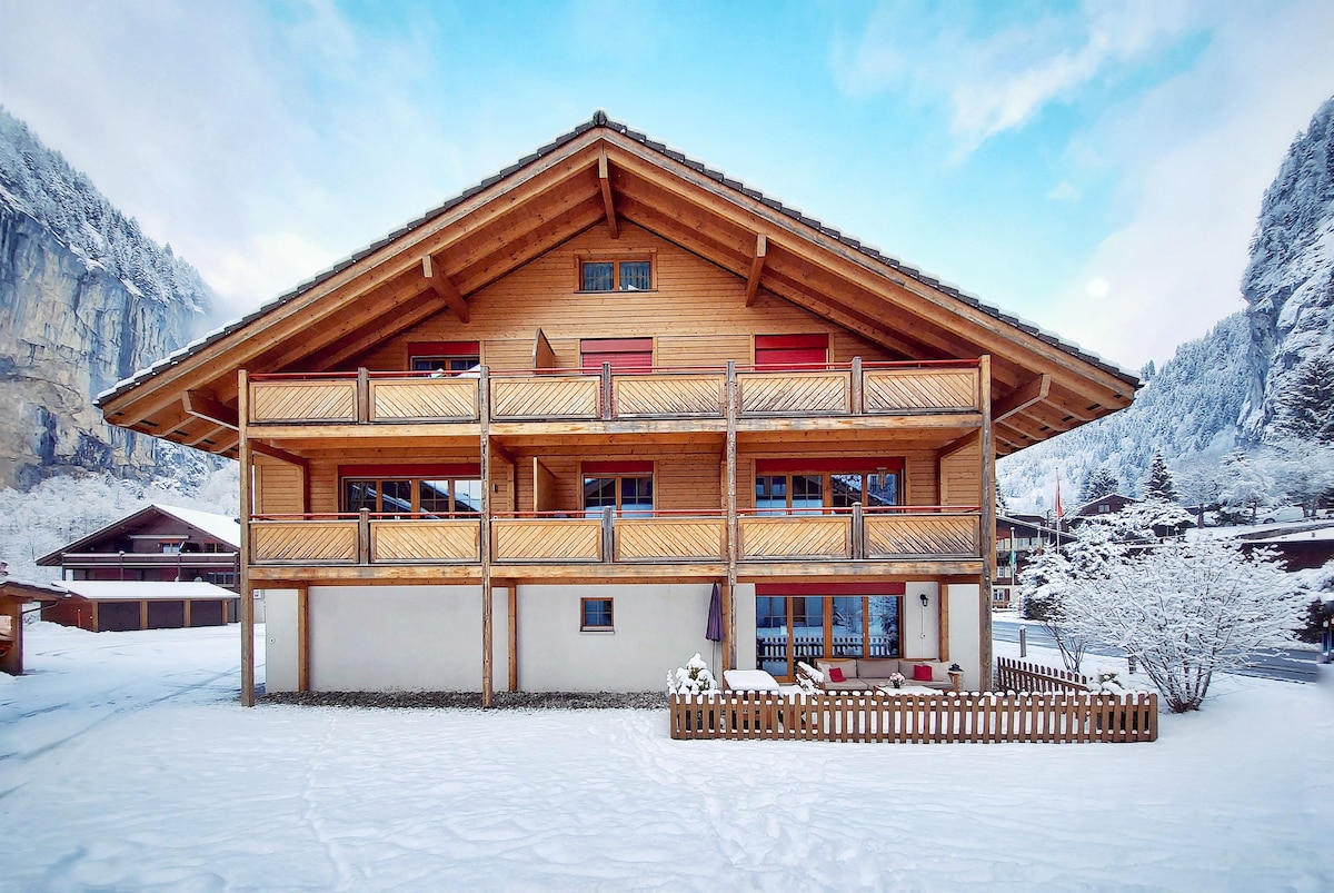 A modern chalet-style building features multiple balconies with wooden railings. The facade is a mix of natural wood and white walls, surrounded by a snowy landscape. The backdrop includes towering mountains under a clear blue sky.