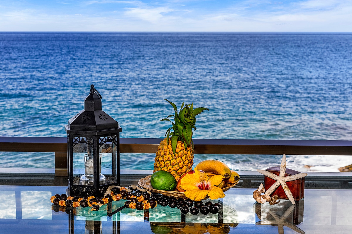 A decorative table is set with a lantern, tropical fruits including a pineapple, and a starfish. The background features an expansive ocean view under a clear blue sky, enhancing the relaxing atmosphere of this oceanfront setting.