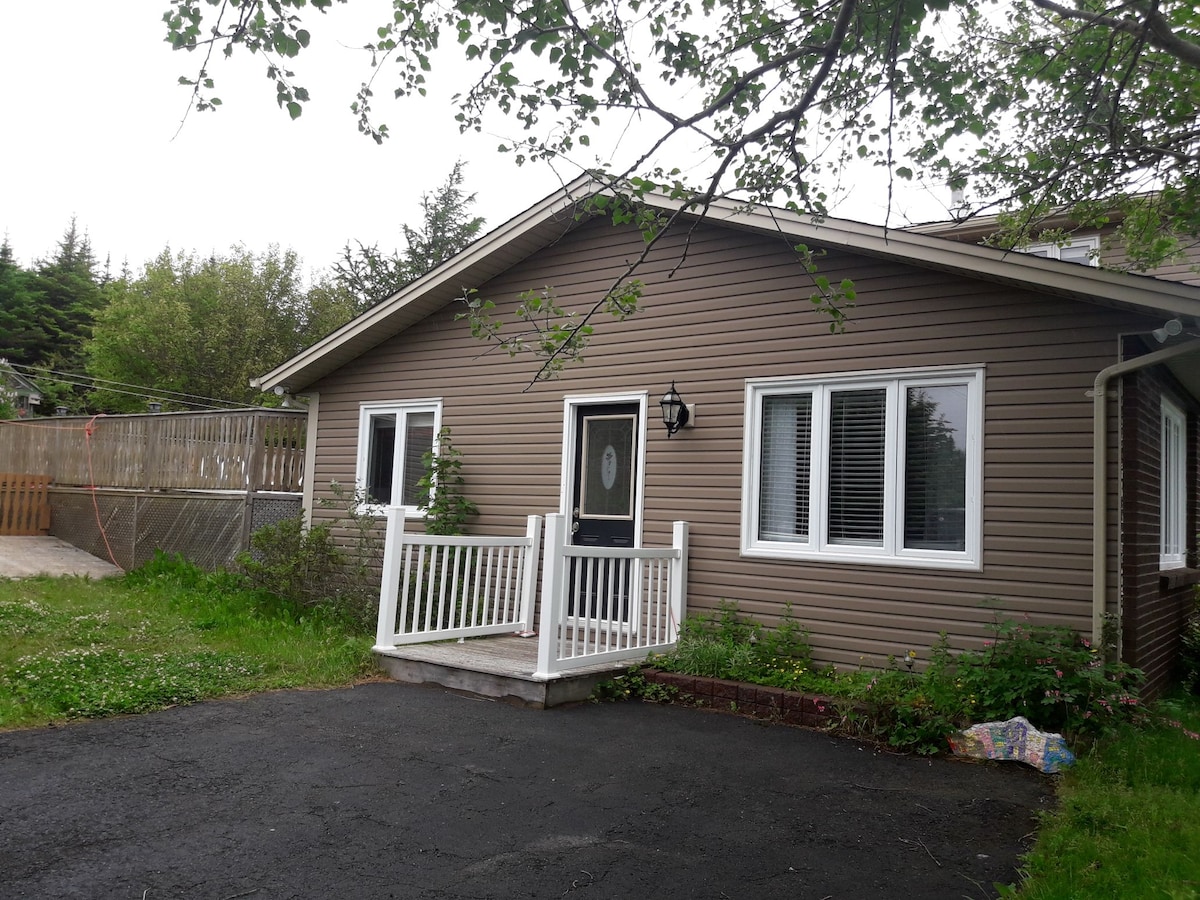 The image displays the exterior of a cozy guest suite, featuring a welcoming entrance with a white railing and front porch. Large windows allow natural light, while a dark asphalt driveway provides convenient access. Surrounding greenery enhances the property's appeal.