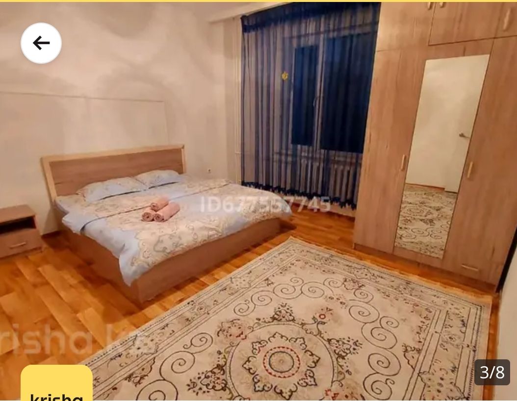 A comfortable bedroom features a spacious bed with light blue bedding and a set of neatly arranged towels. A large mirror is positioned next to the wardrobe, and natural light filters through the striped curtains over the window. A patterned area rug adds warmth to the wooden floor.