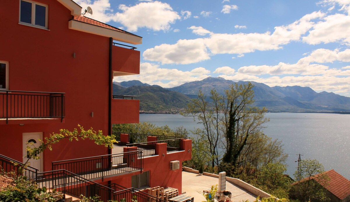 A modern red building sits on a hillside, overlooking a calm body of water. Lush greenery surrounds the structure, with balconies offering scenic views of the mountains and fluffy clouds in a bright blue sky.