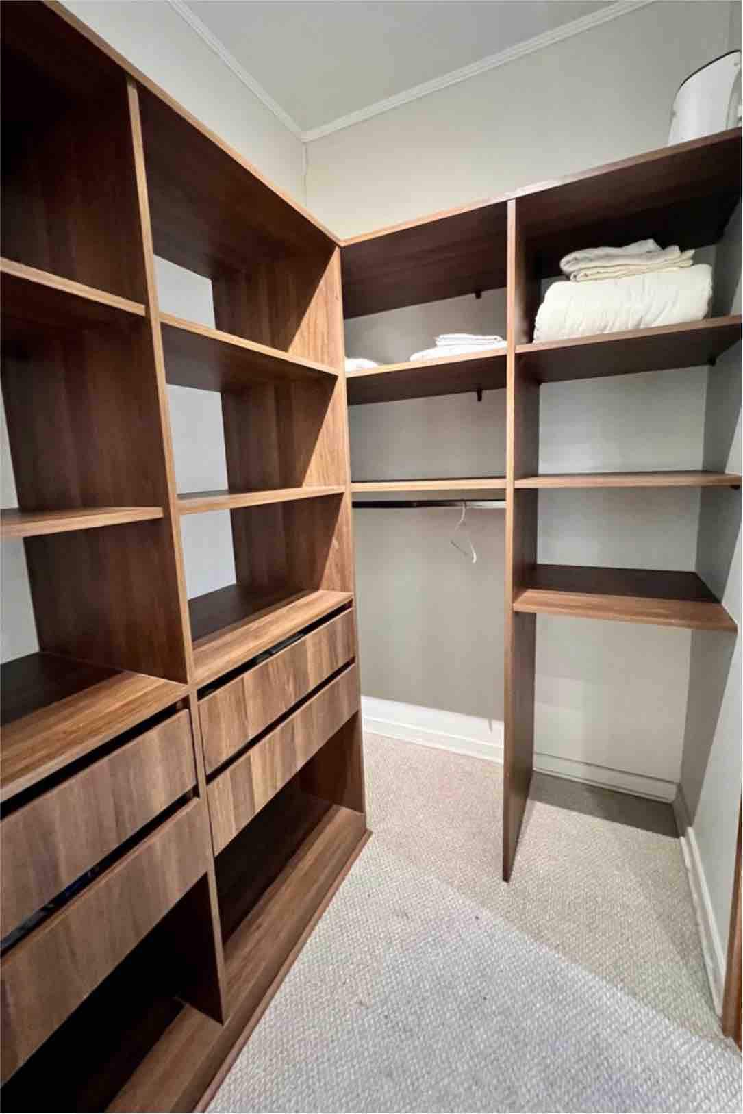 A well-organized closet features wooden shelving and drawers, providing ample storage space. Folded towels are neatly arranged on the top shelf, while open sections offer additional room for belongings. The soft carpeted floor adds warmth to the functional design.